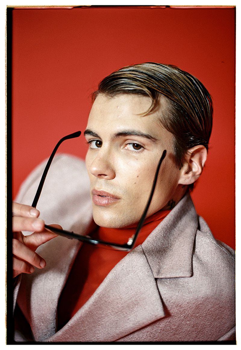 Headshot of a handsome adult male in a blazer holding black-rimmed glasses, red backdrop, and stylish slicked-back hair.