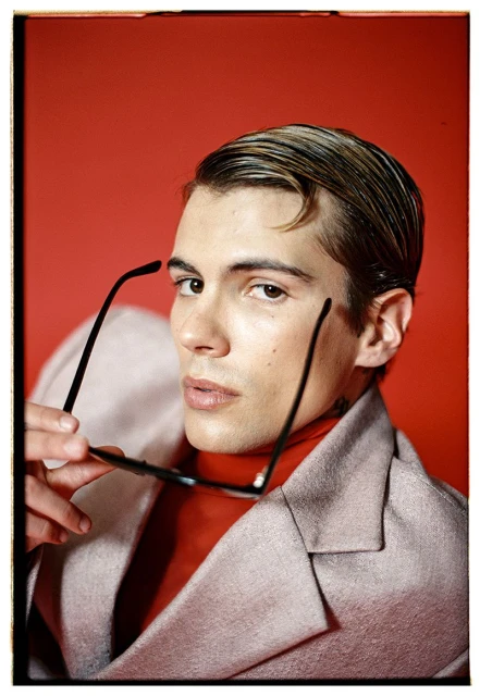 Headshot of a handsome adult male in a blazer holding black-rimmed glasses, red backdrop, and stylish slicked-back hair.
