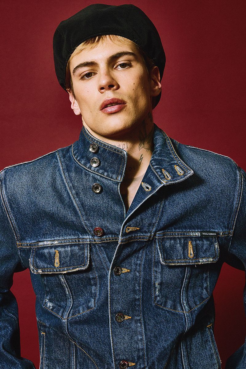 Portrait of a young adult man wearing a denim jacket and a black beret against a red background in a studio setting.