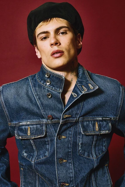 Portrait of a young adult man wearing a denim jacket and a black beret against a red background in a studio setting.