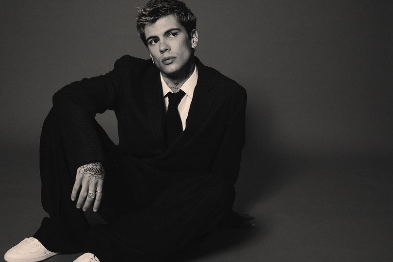 Monochrome portrait of a young adult male in a dark suit, seated against a grey backdrop, showcasing fashion and style in studio lighting.