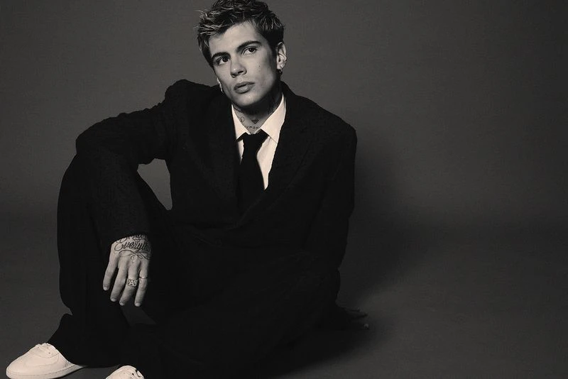 Monochrome portrait of a young adult male in a dark suit, seated against a grey backdrop, showcasing fashion and style in studio lighting.