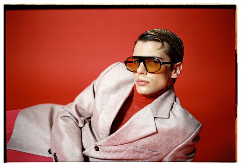 A fair-skinned young adult man in a jacket and yellow sunglasses poses against a red backdrop in a fashion photography studio shot.