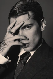 Close-up portrait of a man wearing a dark suit and patterned tie, holding his hand with rings and tattoos partially over his face.