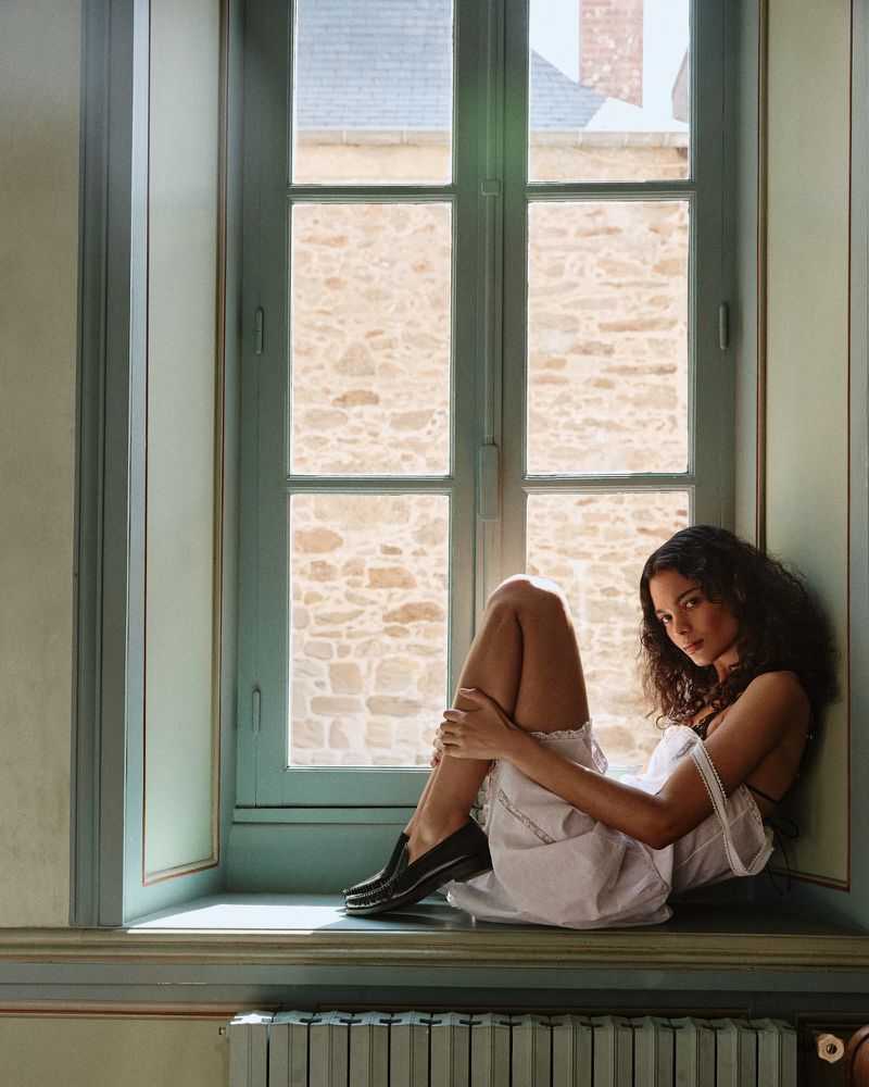 Young woman with dark curly hair wearing a white dress sitting in a window seat, looking toward the camera with a neutral expression.