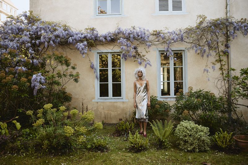 Woman with curly hair stands in a silver dress in a lush green garden with a building adorned with wisteria in the background.