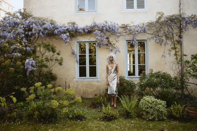 Woman with curly hair stands in a silver dress in a lush green garden with a building adorned with wisteria in the background.