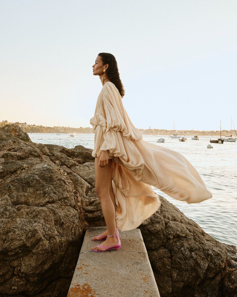 Woman in flowing beige dress stands on rocks overlooking the ocean with sailboats, bathed in soft, diffused light near the coastline.