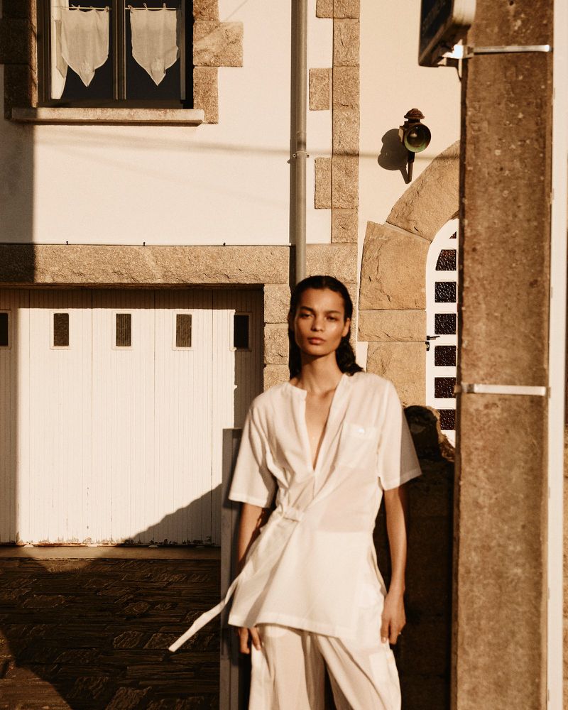 Woman in loose white pantsuit poses against white stone building in golden light. Fashion editorial shot, simple background, minimalist look.