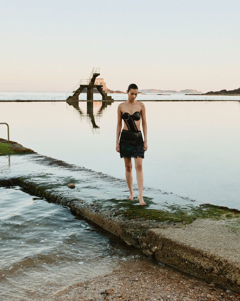 Woman in black dress standing barefoot on concrete dock at water's edge with diving platform at sunset, calm, serene