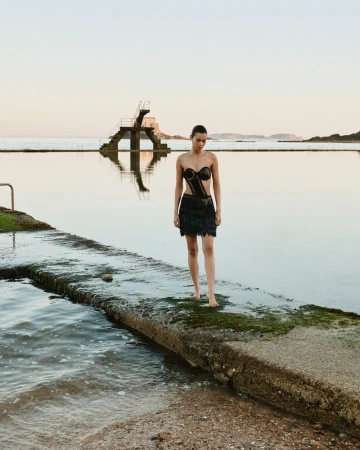 Woman in black dress standing barefoot on concrete dock at water's edge with diving platform at sunset, calm, serene