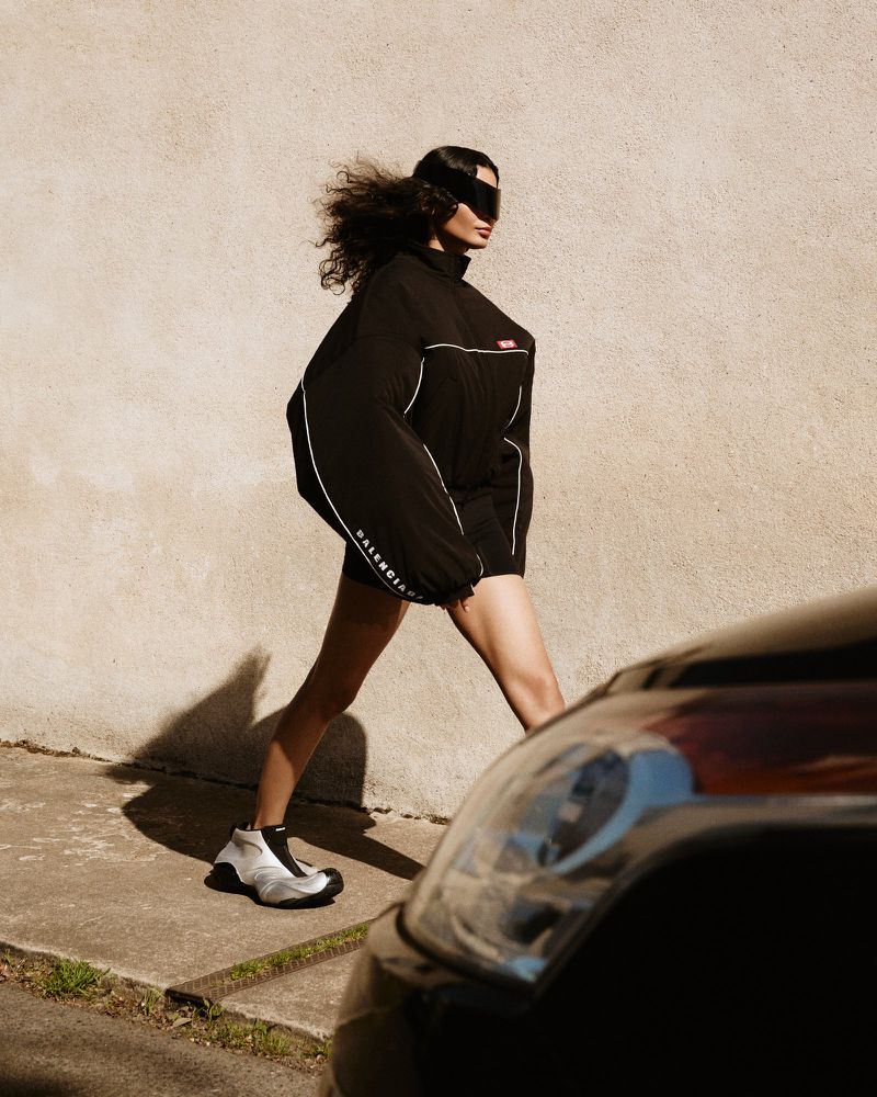 A woman in a black Balenciaga tracksuit walks on a city sidewalk, wearing futuristic sunglasses, with a black car in the foreground.