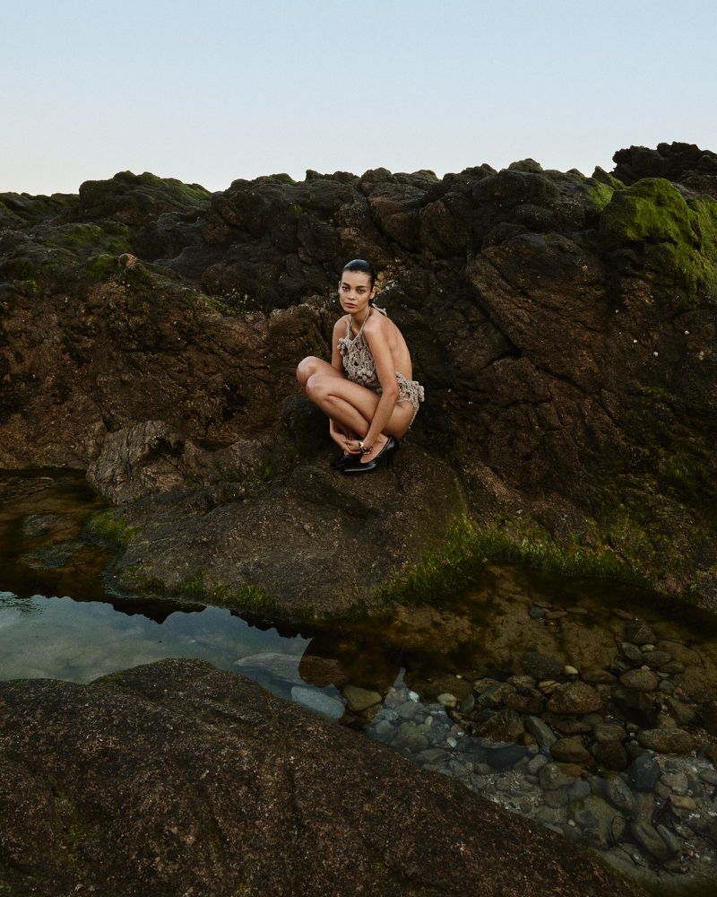 Adult woman in swimwear crouching on rocky beach with moss and water. Ocean scenery under a clear sky in an outdoor setting.