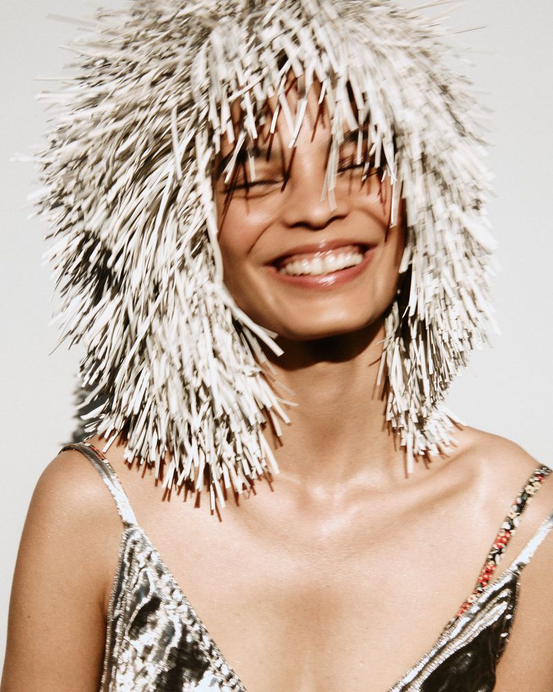 Portrait of a smiling woman with a spiky silver wig and a sequined top against a plain background in a medium shot with soft lighting.