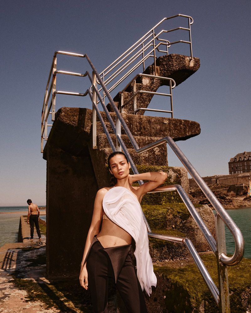 A woman poses in avant-garde fashion next to stairs leading to the ocean, with a figure visible in the background on the beach.