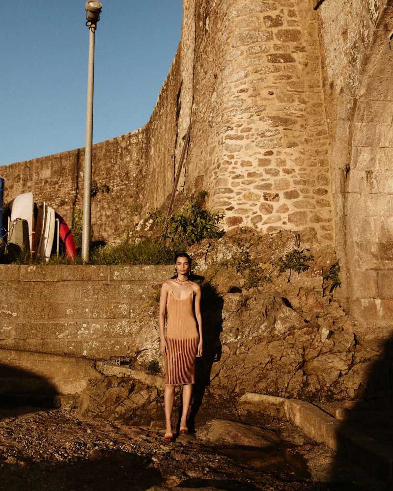 Young woman poses in front of stone wall wearing peach colored tank dress and sandals in an outdoor environment on a sunny day.