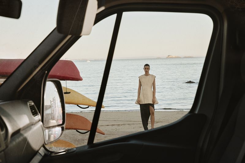Fashion shot on a beach featuring a woman in a dress viewed through a vehicle window with kayaks attached to the vehicle.