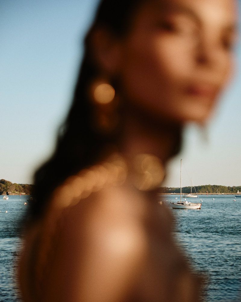 Woman in gold jewelry stands near water with boats under a blue sky in blurred portrait image.