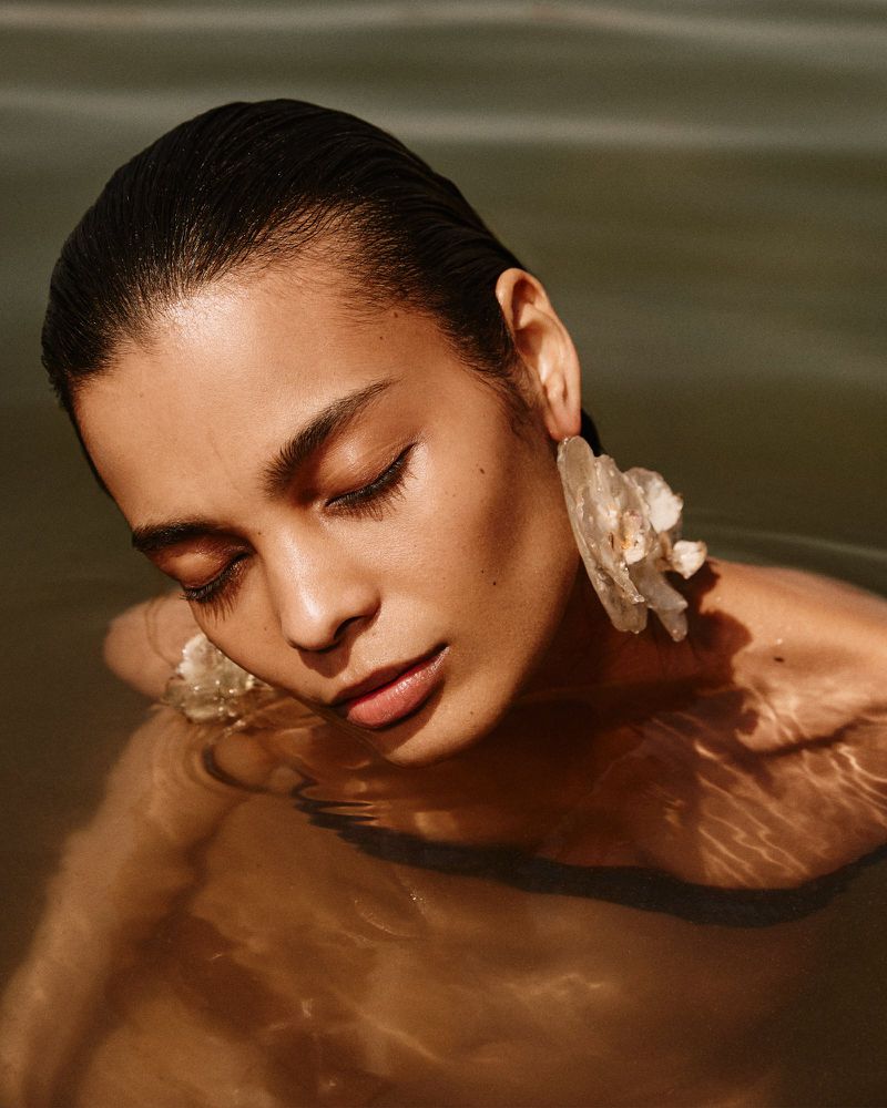 Close-up of a model with wet hair and flower earrings in water, bathed in soft, filtered sunlight, creating a serene and ethereal aesthetic.