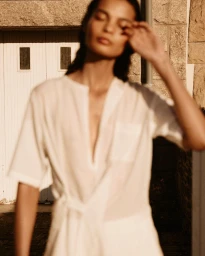 Model in white linen dress standing outdoors against stone building exterior under warm sunlight.