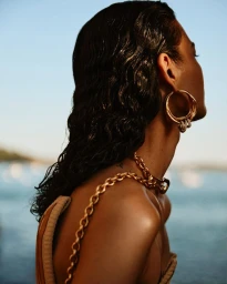Profile view of a model with wet dark hair wearing large gold hoop earrings and a chunky gold chain necklace against an ocean backdrop.