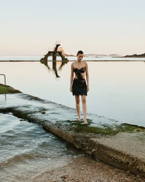 Model standing on a concrete ledge by the water wearing a dark structured top and skirt with a diving platform in the background
