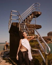 Model leaning against a concrete diving platform outdoors wearing a white draped top and dark trousers.