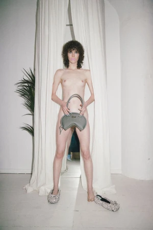 A nude woman with curly hair stands with a grey purse and patterned ballet shoes against a white wall with draped curtains.