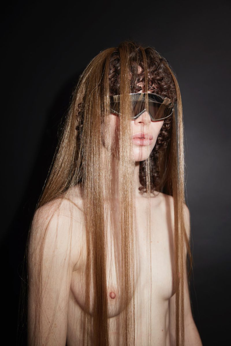 Striking portrait of a topless adult female with wavy brown and blonde hair, wearing reflective sunglasses against a black background.