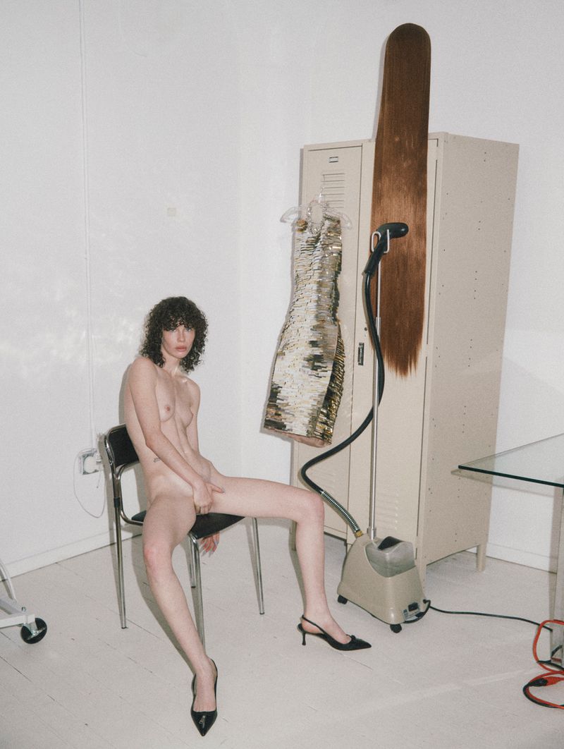 A nude adult female sits in a white studio. In the background are a dress, garment steamer, metal locker, brown wig, chair and glass table.