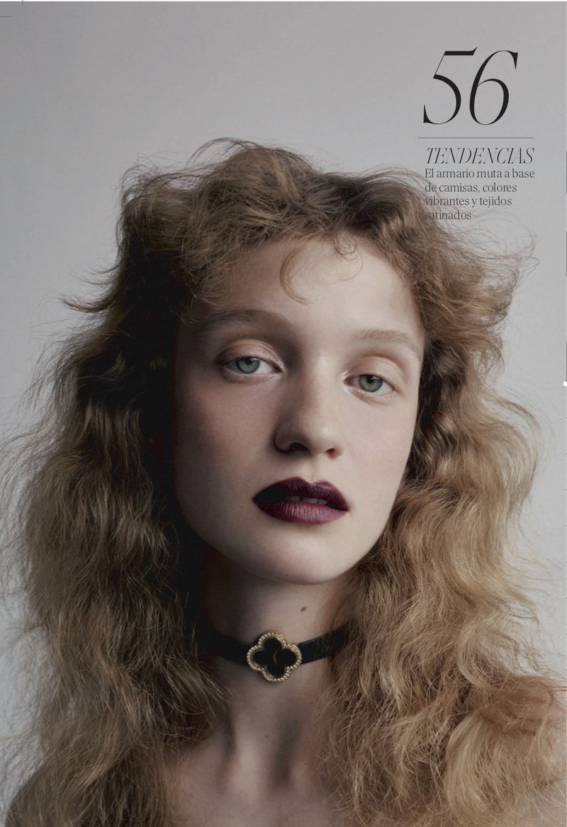 Close-up portrait of a young adult female model with light curly hair and dark lipstick wearing a black and gold choker necklace.