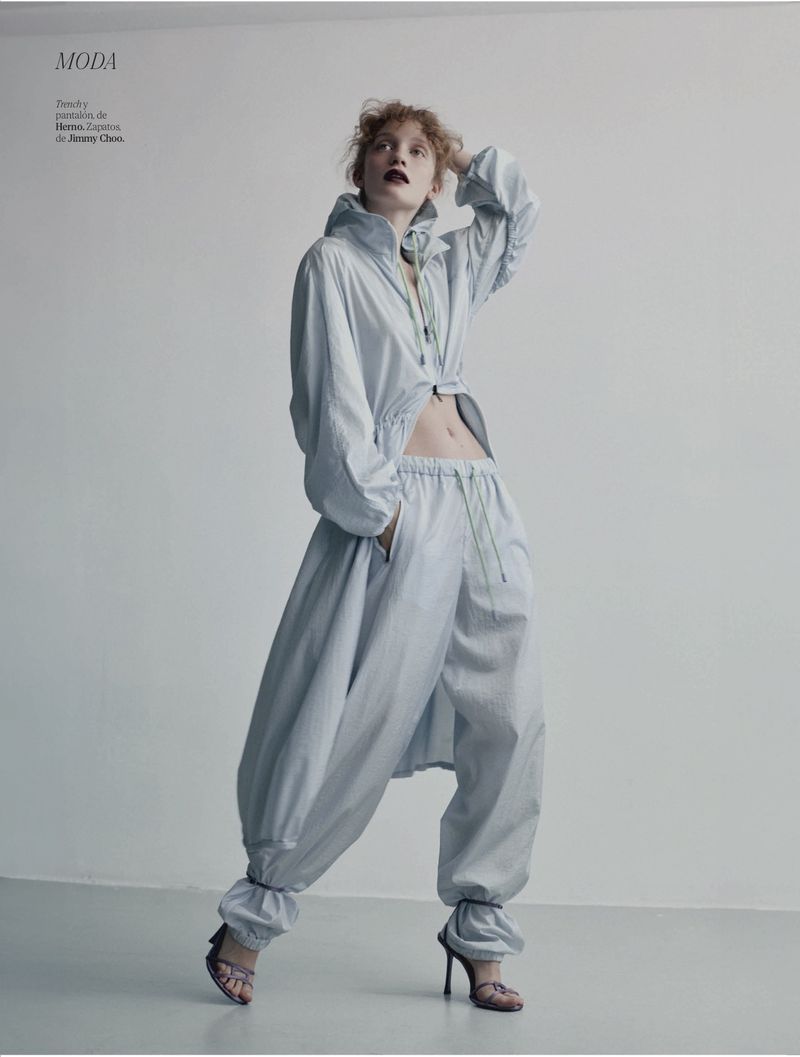 Fashion editorial shot of a woman in a light blue hooded jacket and baggy pants, posing against a plain wall in high heels.