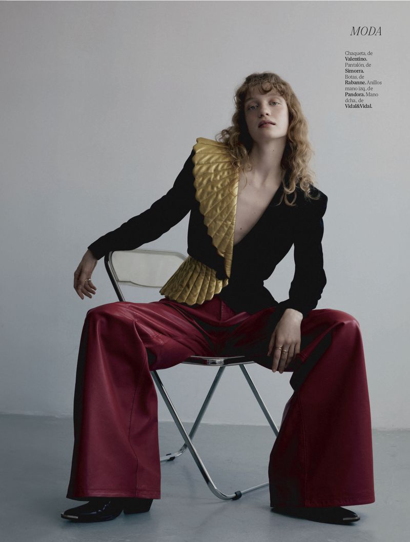 Fashion editorial shot of a model in a black jacket with gold shoulder piece and wide red pants sitting on a clear plastic chair.