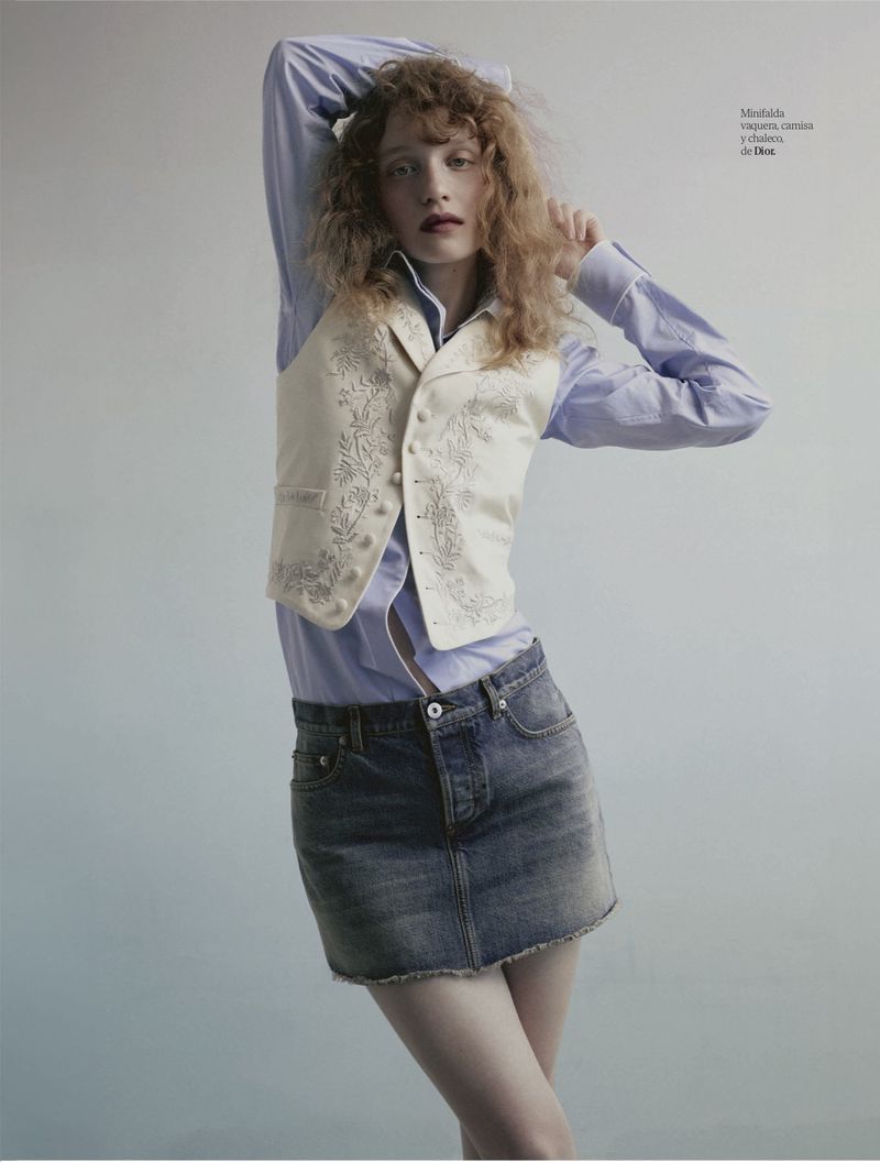 A fair-skinned woman with red hair poses indoors wearing a denim skirt, patterned vest, and light blue button-down shirt with arms overhead.