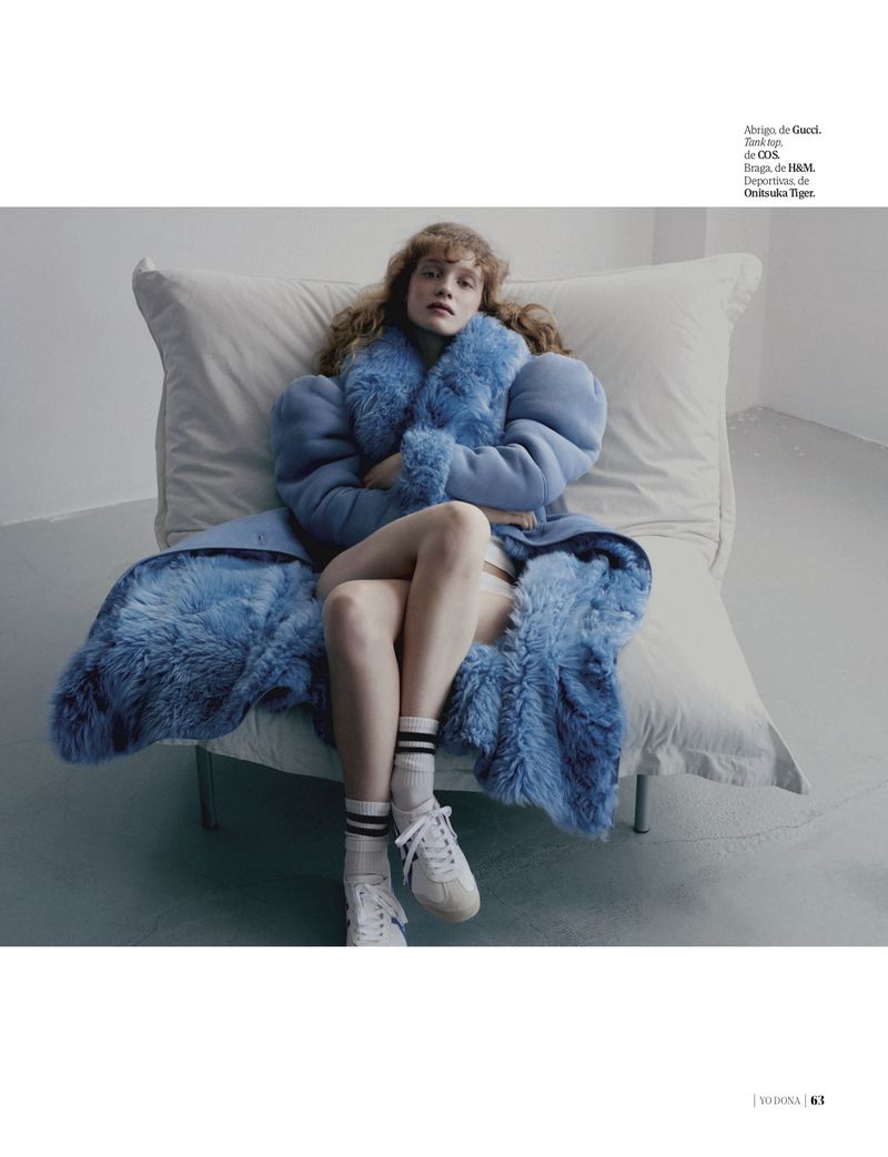 A woman with wavy red hair wearing a blue fur-trimmed coat and white sneakers sits on a bed with white pillows against a grey background.