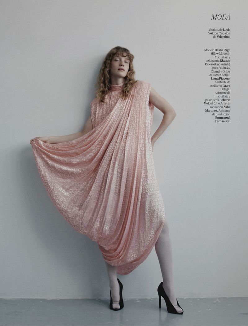 Fashion editorial featuring a woman with curly hair wearing a sparkly pink gown and black high heels posing in a studio against a white wall.