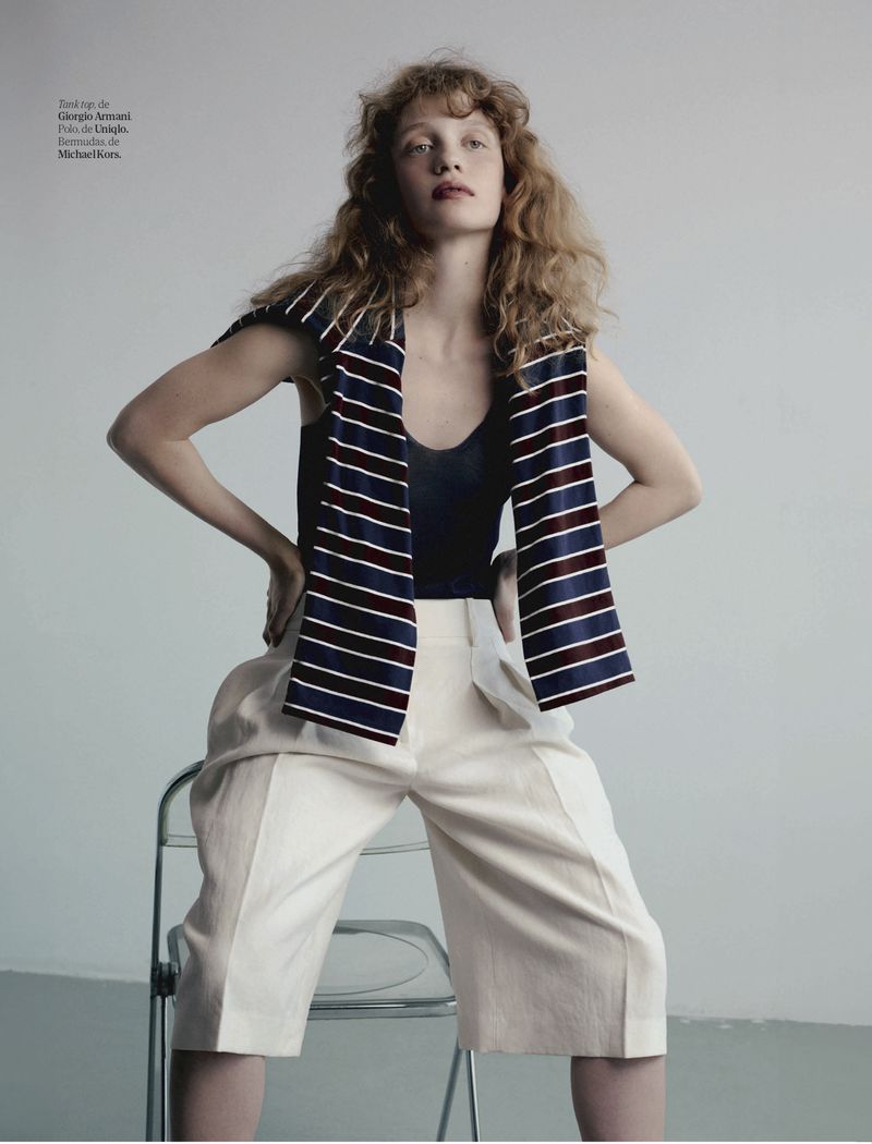 Fashion editorial shot of a stylish young woman in navy top, white pants, and striped scarf, posing on a clear chair in a studio.