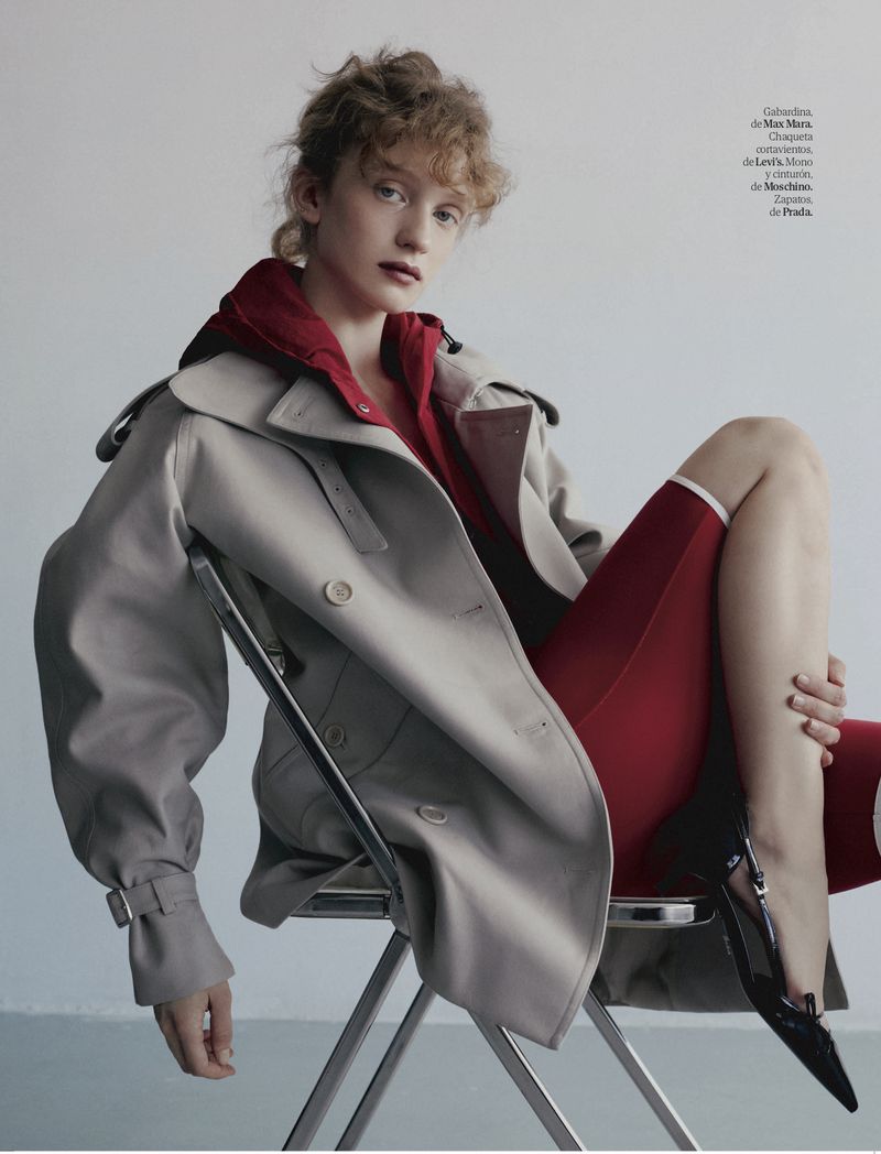 Fashion editorial shot of a female model wearing a beige trench coat and red apparel, sitting on a chair in a minimalist setting.