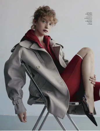 Fashion editorial shot of a female model wearing a beige trench coat and red apparel, sitting on a chair in a minimalist setting.