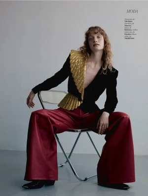 Model seated on a chair wearing a black jacket with gold pleated ruffles and wide red leather trousers against a grey wall