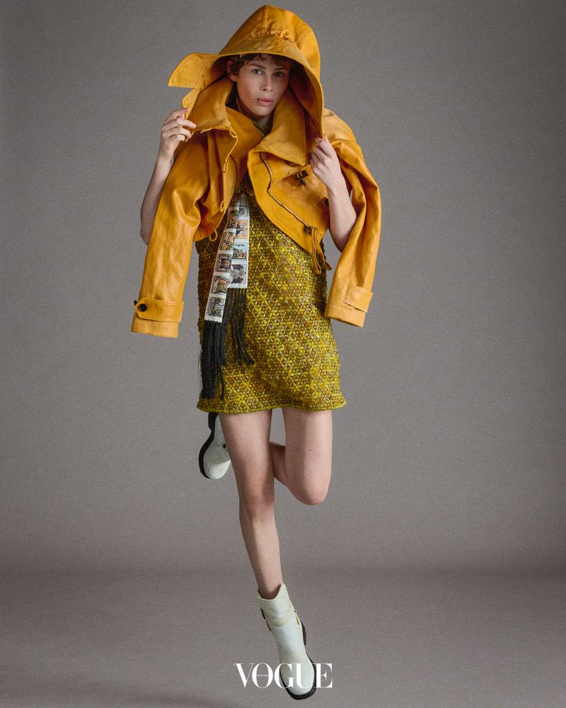 Fashion editorial shot of a woman in a yellow dress and jacket with white boots, posing in a studio with a gray background.