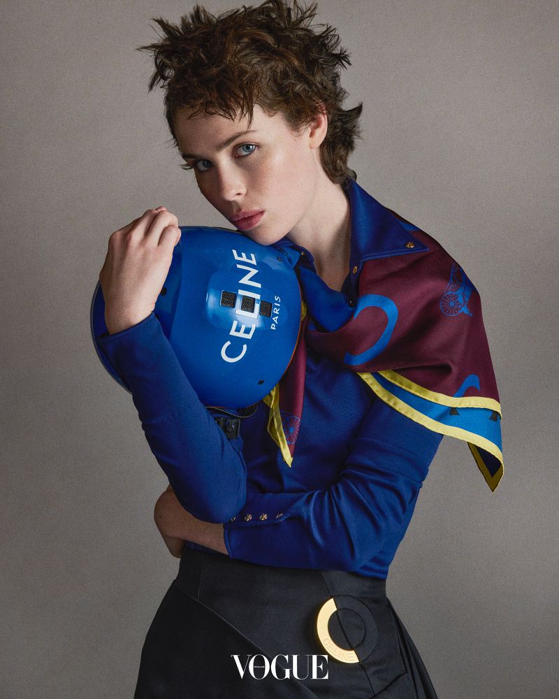 An adult woman poses indoors, wearing a blue top, black skirt, and scarf. She holds a blue helmet with the word 'Celine' printed on it.