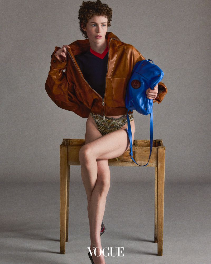 Fashion shot of a model sitting on a wooden table, wearing a brown jacket, beaded bottoms, and a blue purse in a grey photo studio.
