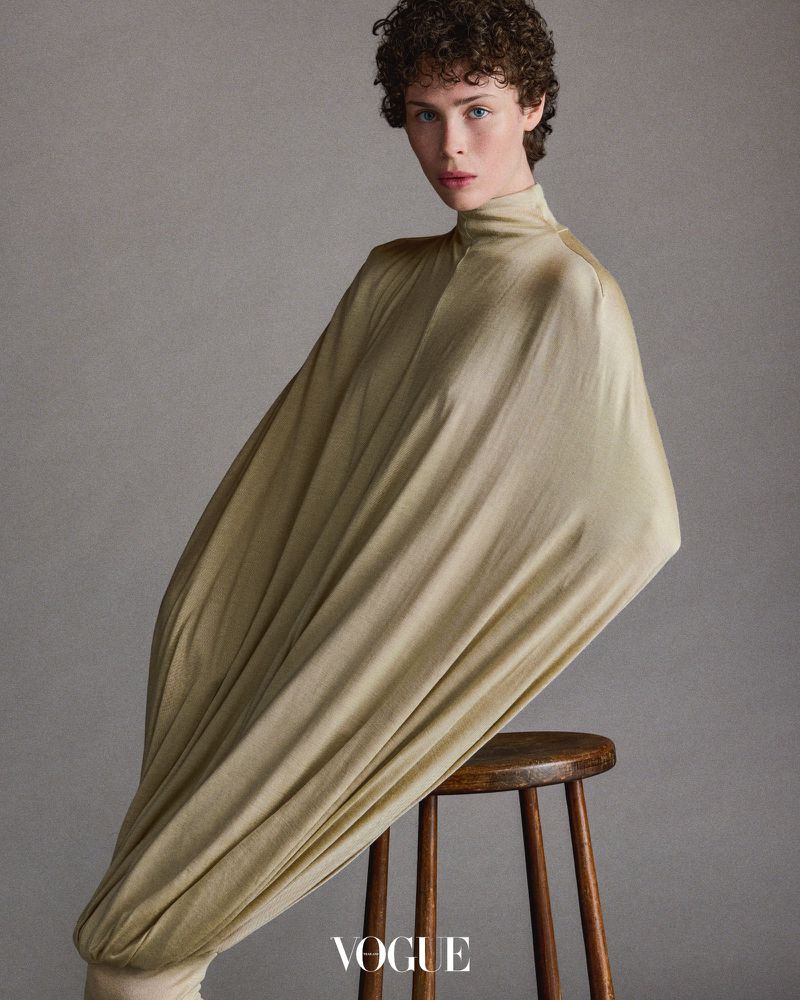 Fashion shot of a young adult woman with curly hair posing in a draped gold fabric dress on a wooden stool in a neutral studio.