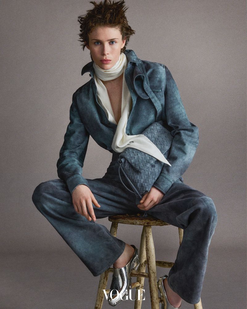 Woman in blue suit with patterned purse, sitting on wooden stool in studio. Fashion editorial image for Vogue with neutral backdrop.