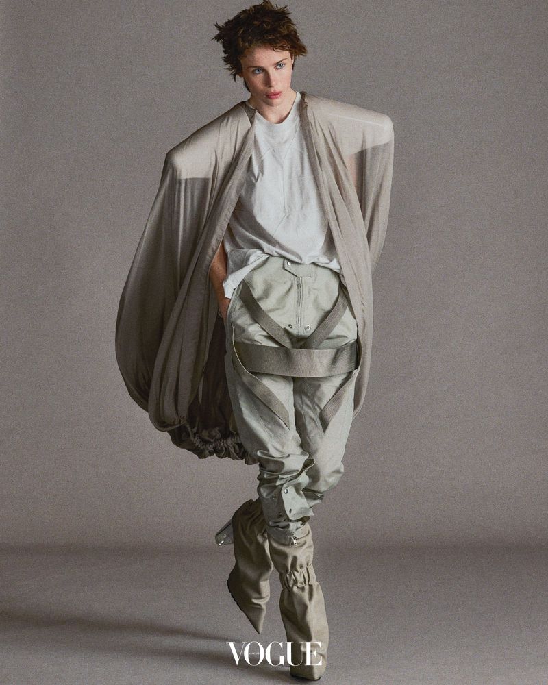 Fashion shot of a person in light-colored, layered clothing, including baggy trousers and an oversized cardigan, with studio lighting.