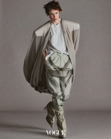 Fashion shot of a person in light-colored, layered clothing, including baggy trousers and an oversized cardigan, with studio lighting.