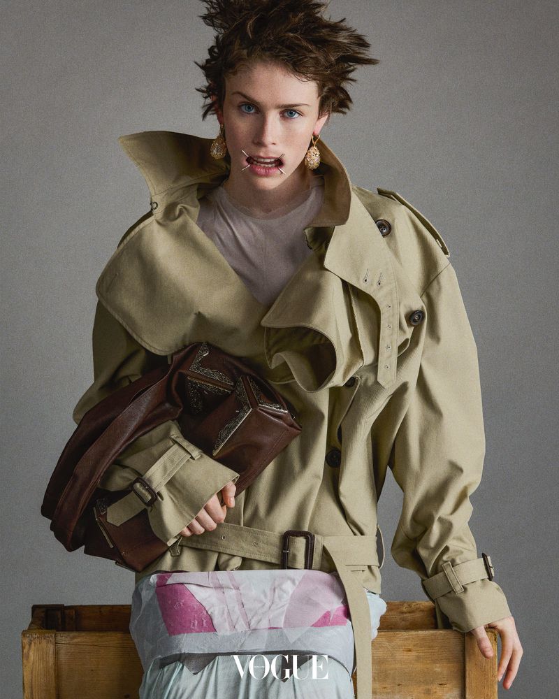 Woman in a trench coat holding a brown leather bag posing in a studio against a neutral backdrop, with the Vogue logo at the bottom.