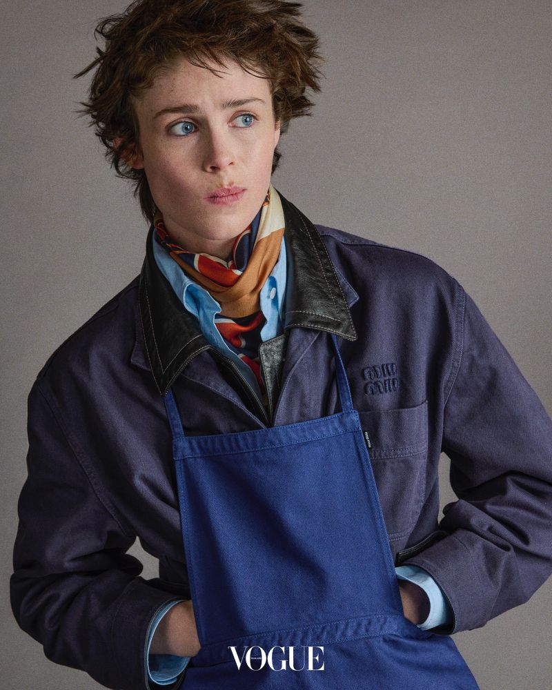 Androgynous young adult woman wearing a blue denim jacket and apron, shot in a studio with muted tones for a fashion editorial.
