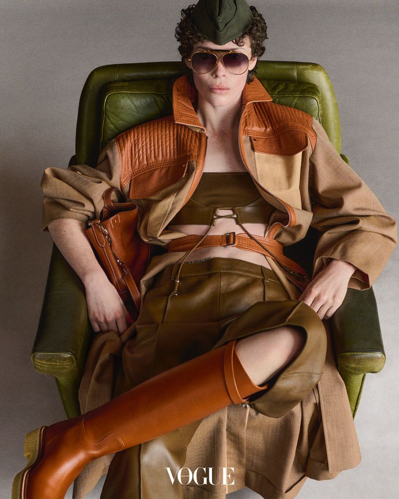 A model poses in an olive green chair, wearing a jacket, top, trousers, boots, and sunglasses, with a small handbag, shot in a studio.